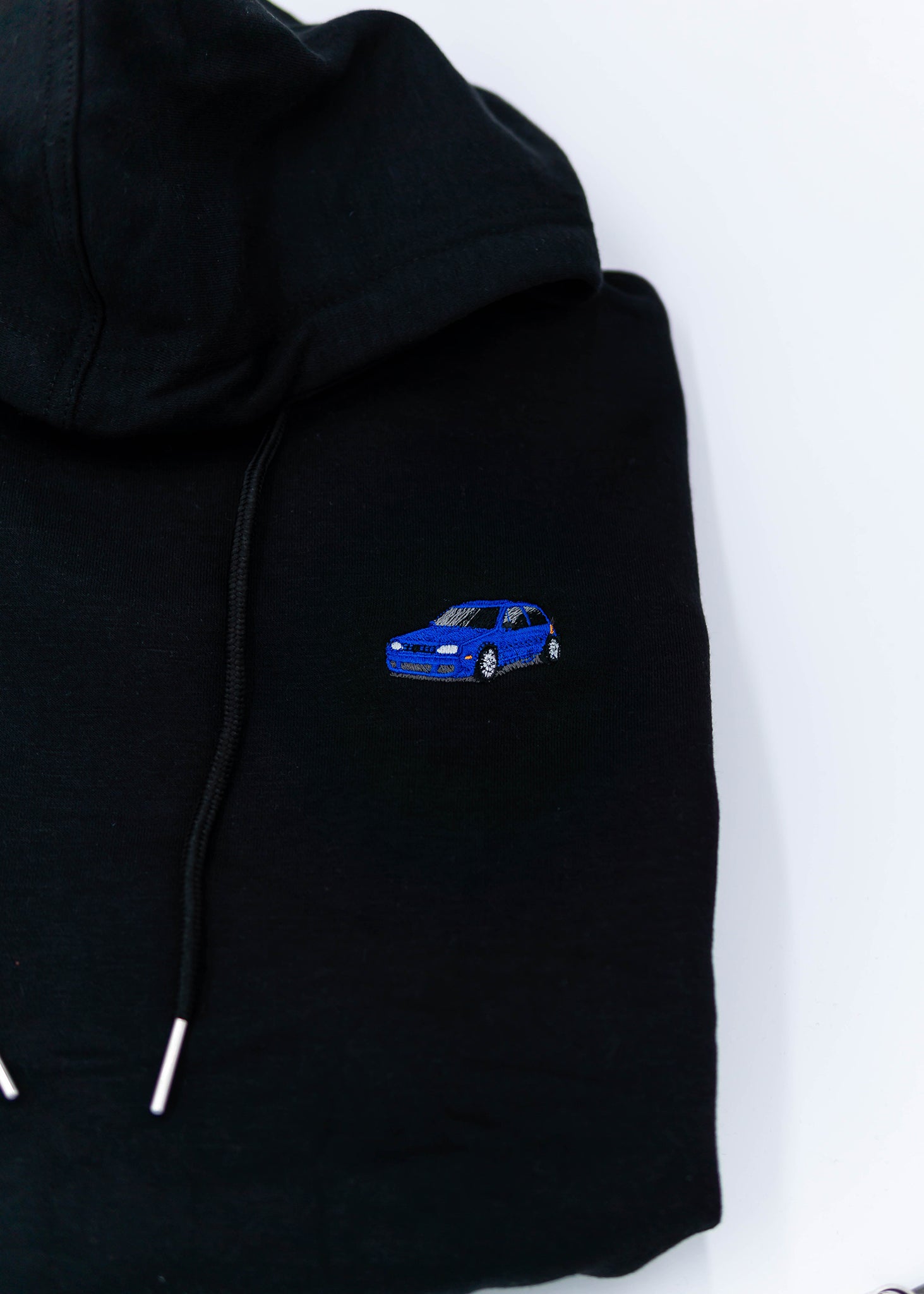 A black Mk4 Golf R32 cropped hoodie for women. Photo is a close up view of the cropped sweater with an embroidered blue Mk4 Golf R32. Fabric composition is cotton. The material is soft, stretchy, and non-transparent. The style of this crop hoodie is long sleeve, crewneck with a hood, hooded, with embroidery on the chest.